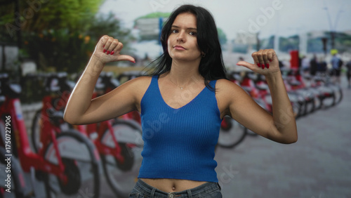 Hispanic young woman pointing both thumbs to herself beside a line of red rental bikes on a busy city street; pride.