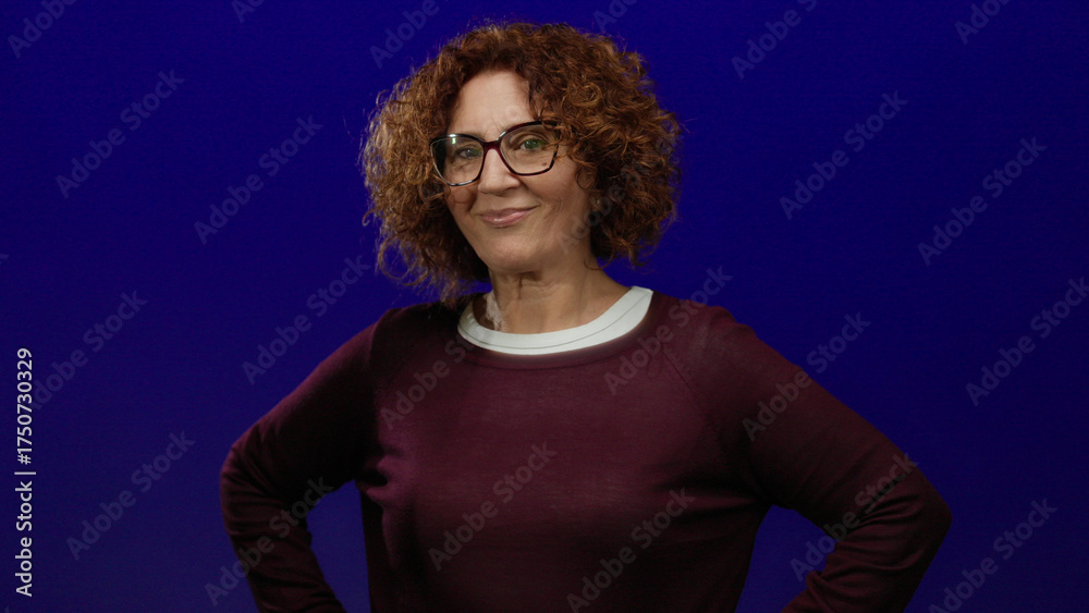 Naklejka premium Woman with curly hair smiling confidently against an isolated blue background wearing glasses and casual attire, embodying positivity and friendliness in a studio setting.