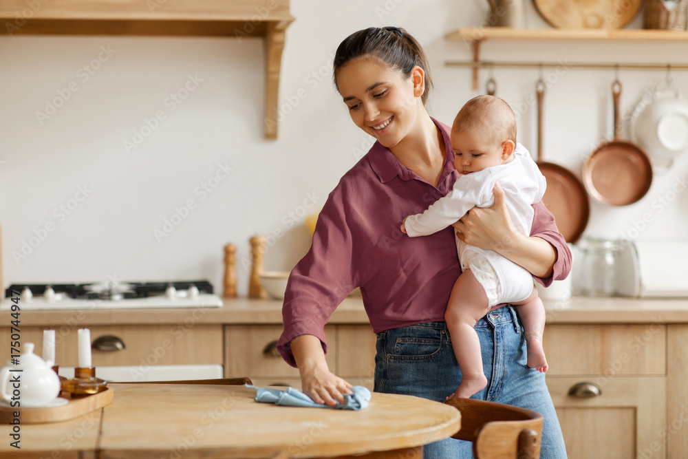 Fototapeta premium Parenthood and multitasking. Mother holding little baby and cleaning table in kitchen, doing house chores, free space