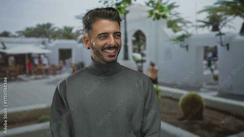 Fototapeta premium Hispanic man in gray turtleneck with beard smiling broadly under sunlight on busy street; optimism leisure.