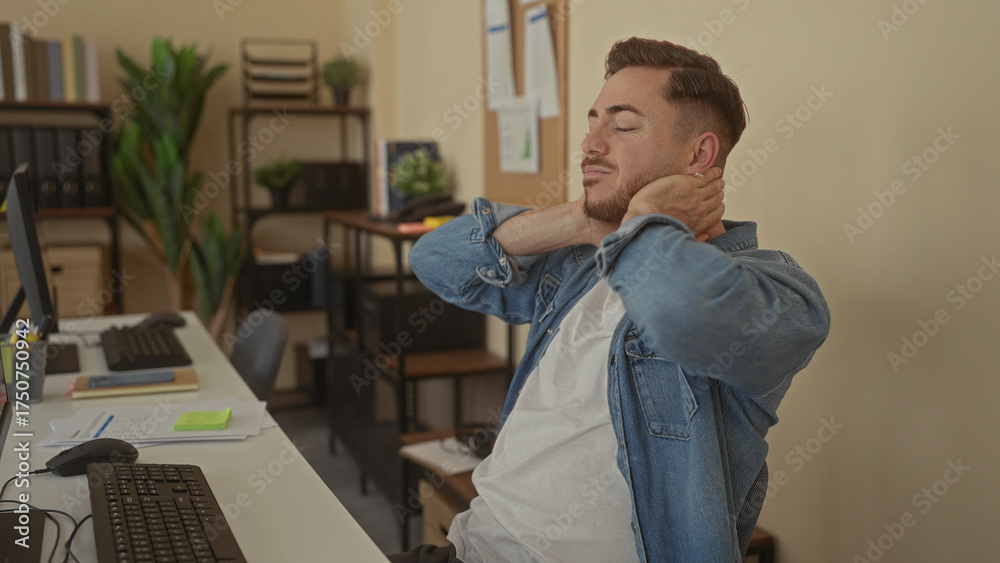 Fototapeta premium Young caucasian man with beard in office stretches neck wearing casual denim surrounded by plants and office equipment, creating a relaxed indoor work environment.