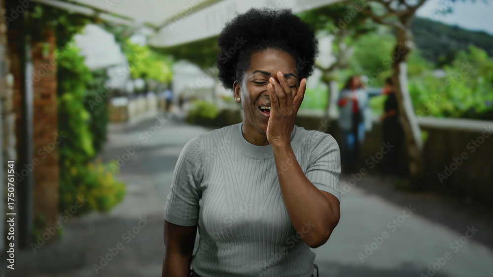 Fototapeta premium Woman laughing with hand on face outdoors on a city street with greenery in the background, capturing a candid moment of joy.
