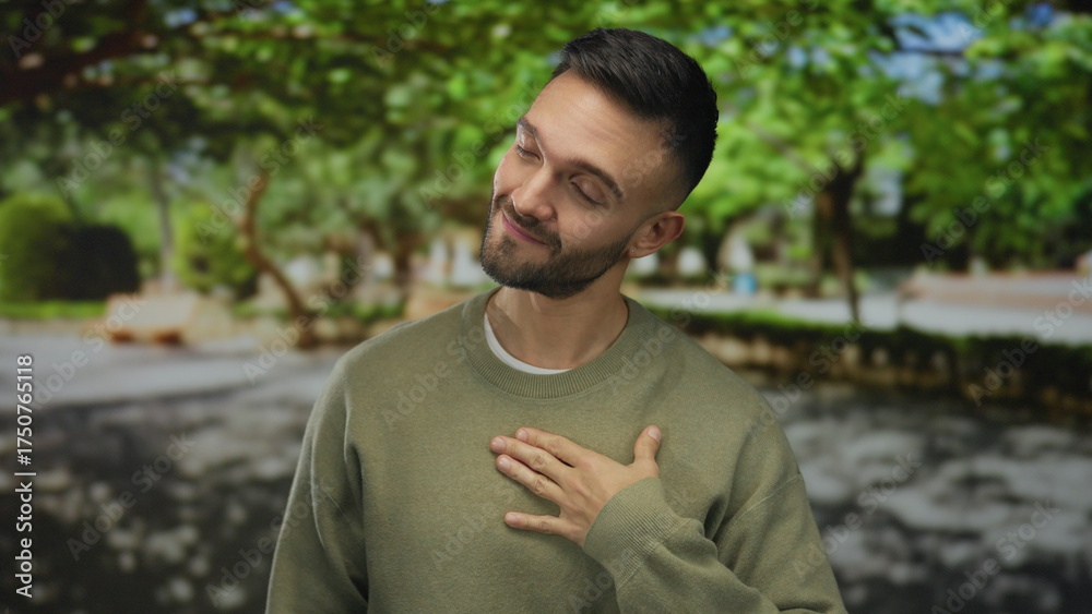 Fototapeta premium Young hispanic man standing outdoors in a green park, smiling warmly, hand on chest, surrounded by lush trees, exuding calm and peaceful vibes, dressed casually.