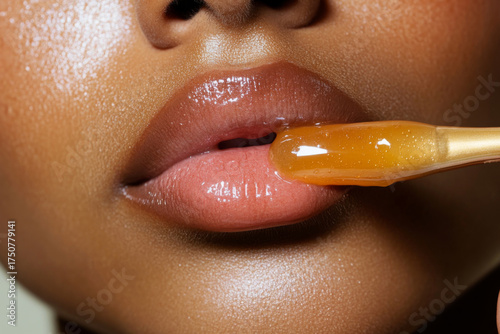 Close-up of glossy lips with a honey-like substance being applied using a wooden stick, highlighting texture and shine.