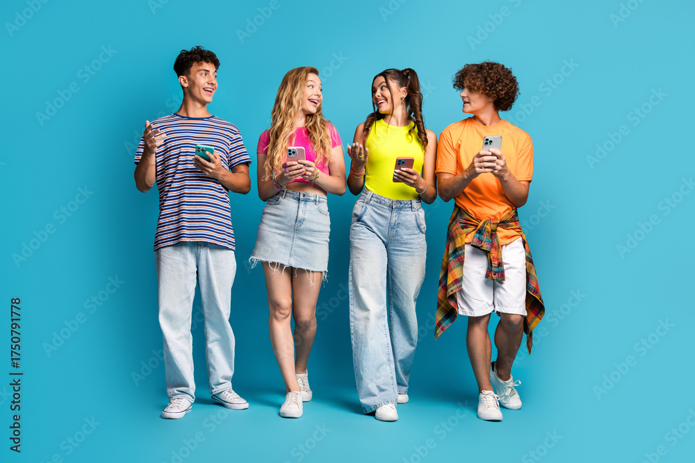 Obraz premium Playful group of friends chatting and texting on phones in bright casual outfits against a vibrant blue backdrop