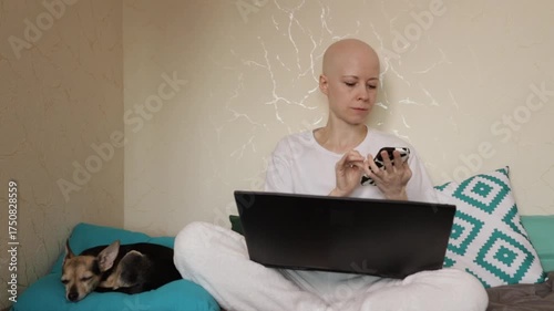 Female cancer patient with alopecia in pajamas smiling while texting on smartphone, cozy bedroom scene with pet dog for comfort