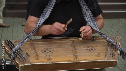 A street musician in a black T-shirt plays the ancient traditional national instrument, the dulcimer, with professional virtuosity and ease using wooden sticks, while spectators walk behind him.