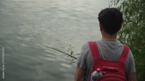 A woman with short dark hair stands shore city lake near a willow tree looks attentively and intently float fishing rod while fishing, holding large red backpack back, calm dark water