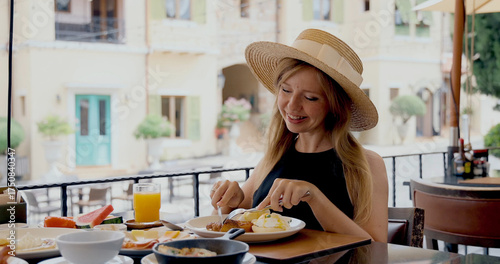 Woman dining in outdoor cafe enjoying breakfast, wearing sunhat in charming European village. Concept of travel, leisure, summer, lifestyle.