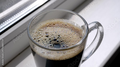 A transparent mug of freshly brewed black coffee with rich foam sits on a bright white windowsill, catching the morning sunlight. Ideal for cozy morning, breakfast, or relaxation themes.