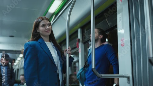 Caucasian woman with long hair wearing blue coat standing on city bus holding pole. Female smiling softly while waiting for stop. Man in blue sweater with backpack standing near door after work.
