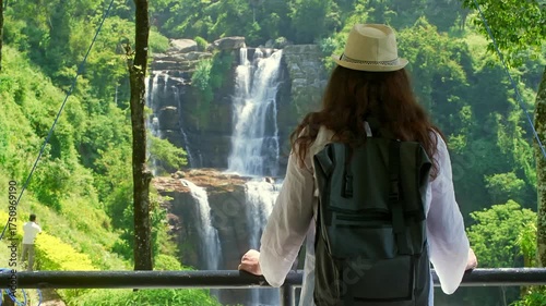 Young female traveler wearing backpack and sun hat standing near powerful waterfall cascading through lush green tropical valley, enjoying scenic natural landscape during adventurous hiking trip