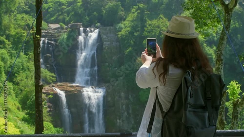 Young female traveler wearing backpack and hat capturing scenic waterfall views in lush green valley during summer adventure, documenting journey with smartphone photography