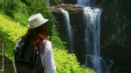 Redhead traveler wearing white hat and carrying backpack standing near cascading waterfall, surrounded by lush green valley with dramatic rocky landscape and verdant vegetation