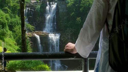 Backpack wearing traveler leaning against railing, absorbing panoramic waterfall scenery within lush tropical forest surroundings, embodying peaceful nature exploration