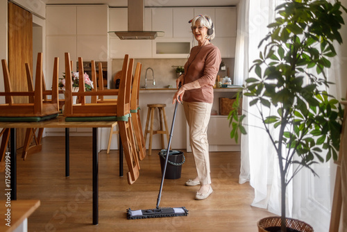Elderly woman wearing headphones singing while mopping parquet kitchen floor with mop and bucket at home