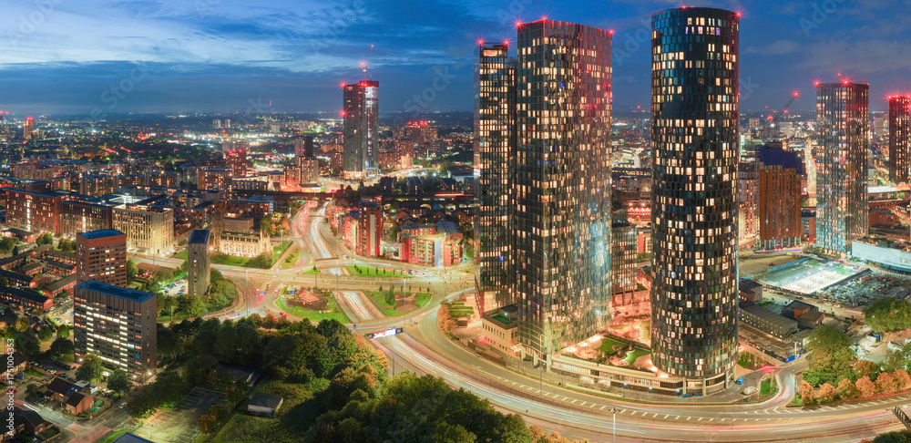 Naklejka premium Blue-hour panorama of Manchester skyline with Deansgate Towers and glowing traffic trails at Mancunian Way roundabout. 