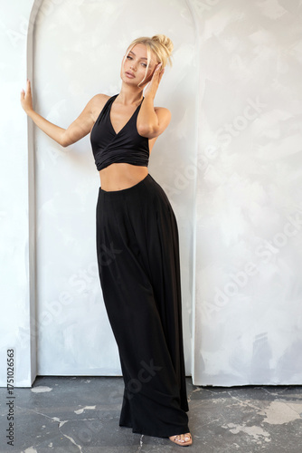 Confident blonde woman posing in a minimalist studio, dressed in a black halter top and wide-leg pants. Fashion-forward, urban look with subtle sensuality. Ideal for fashion