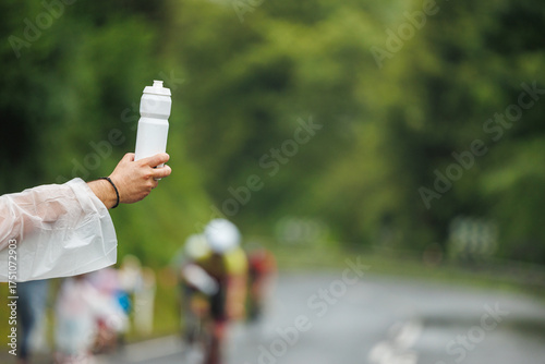 Close-up of a hand holding out a white water bottle to passing cyclists, captured in natural style with a blurred forest background, concept of hydration support