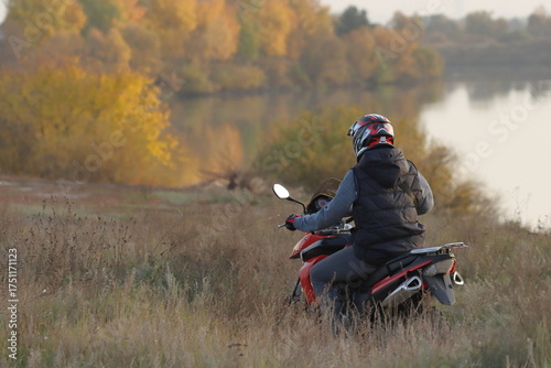 A man on a motorcycle rides along a river on an autumn evening against the backdrop of yellow leaves on the trees along the bank
