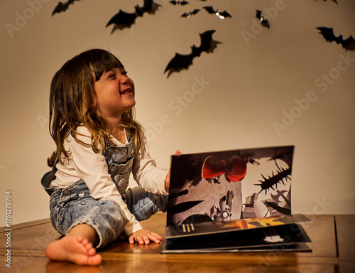 Charming child or smiling girl in overalls reads a Halloween book under warm lighting, with bat wall decorations creating a cozy, spooky ambiance. Autumn, storytelling, and spooky festive concept