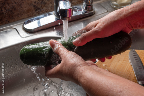 Manipulacion de alimentos, manejo de comida en la cocina, manos agarrando un pepino para lavarlo con agua de la llave en la tarja de una cocina, sobre una tabla de cortar