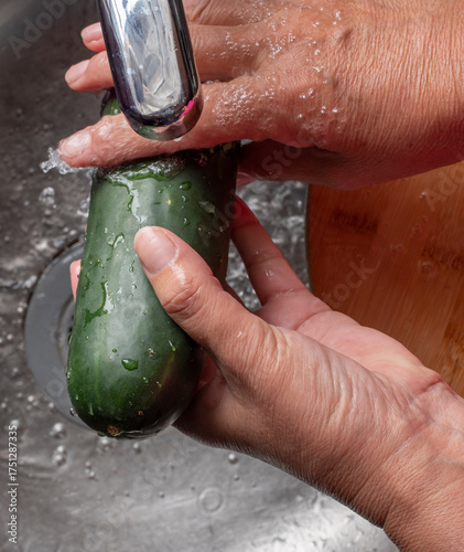 Manipulacion de alimentos, manejo de comida en la cocina, manos agarrando y lavando pepino con chorro de agua de la llave con una tabla de cortar al fondo.