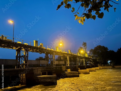 A long bridge with lights at night over a river, with trees in the background and a clear sky