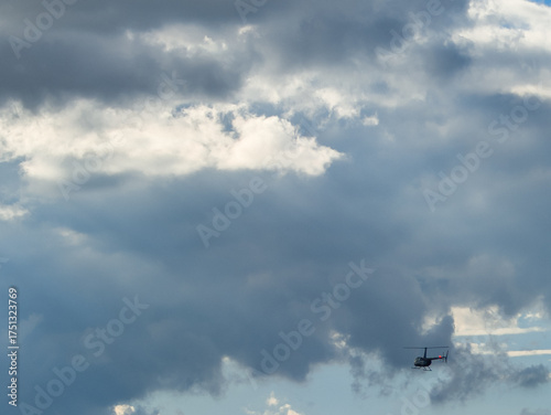 Small black helicopter maneuvering against a background of dense overcast cumulus clouds with soft sunlight filtering through the grey sky on a crisp and windy autumn day with deep shadows
