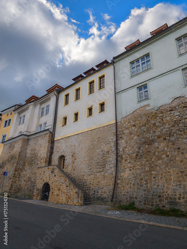 Perspective view of a tall ancient stone fortification wall supporting colorful residential houses with a small wooden door and stone stairs under a bright blue sky with white clouds.