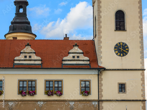 Close up of a vintage clock tower with golden roman numerals on a cream facade and a red tiled roof with floral window boxes under a bright blue sky.
