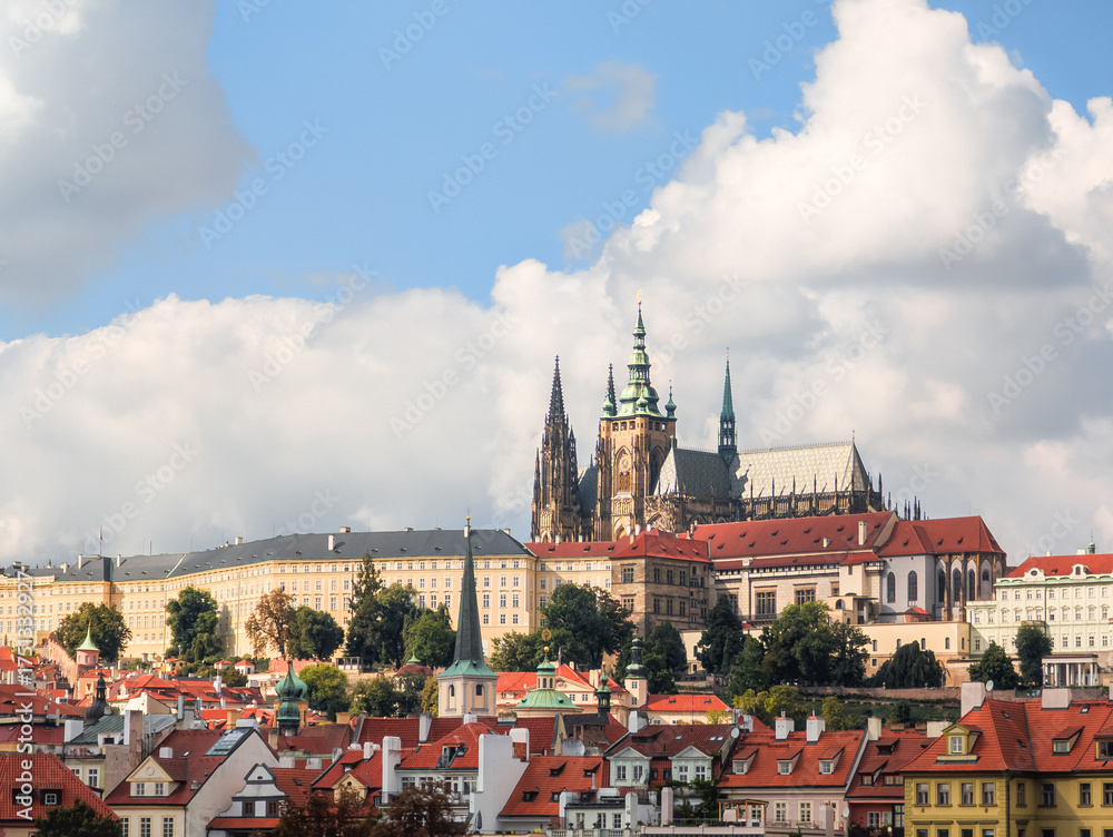 Obraz premium Majestic gothic cathedral rising above historic red rooftops under a bright blue sky with fluffy white clouds during a sunny autumn afternoon showing intricate stone architecture and towers.