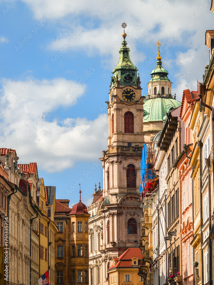 Obraz premium Vertical view of an ornate baroque church tower with a green copper dome and a large black clock face standing against a blue sky with soft white clouds and sunlit facades.