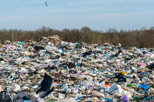 Expansive outdoor landfill site overflowing with assorted waste, including plastics and debris, beneath a bright sky with distant trees