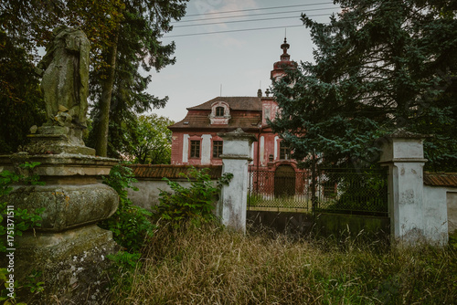 Historic stone sculpture standing in front of an old pink mansion with a clock tower under a soft evening sky in a rural landscape at sunset.