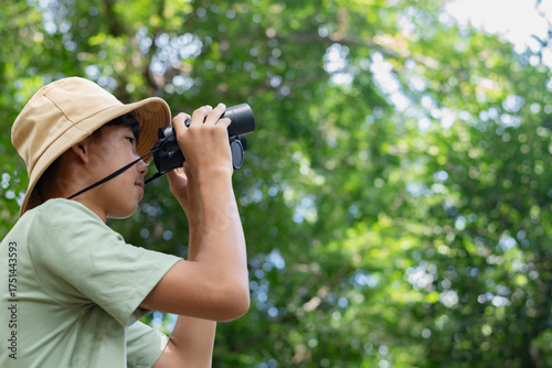 Fotografie A young naturalist looks high into the treetops using his binoculars, with a wide expanse of green foliage filling the background
