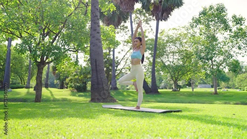 Asian woman standing yoga poses meditating in public park, stand on green grass with trees in the background on a sunny day. Concept of mindfulness, self-care, and outdoor relaxation
