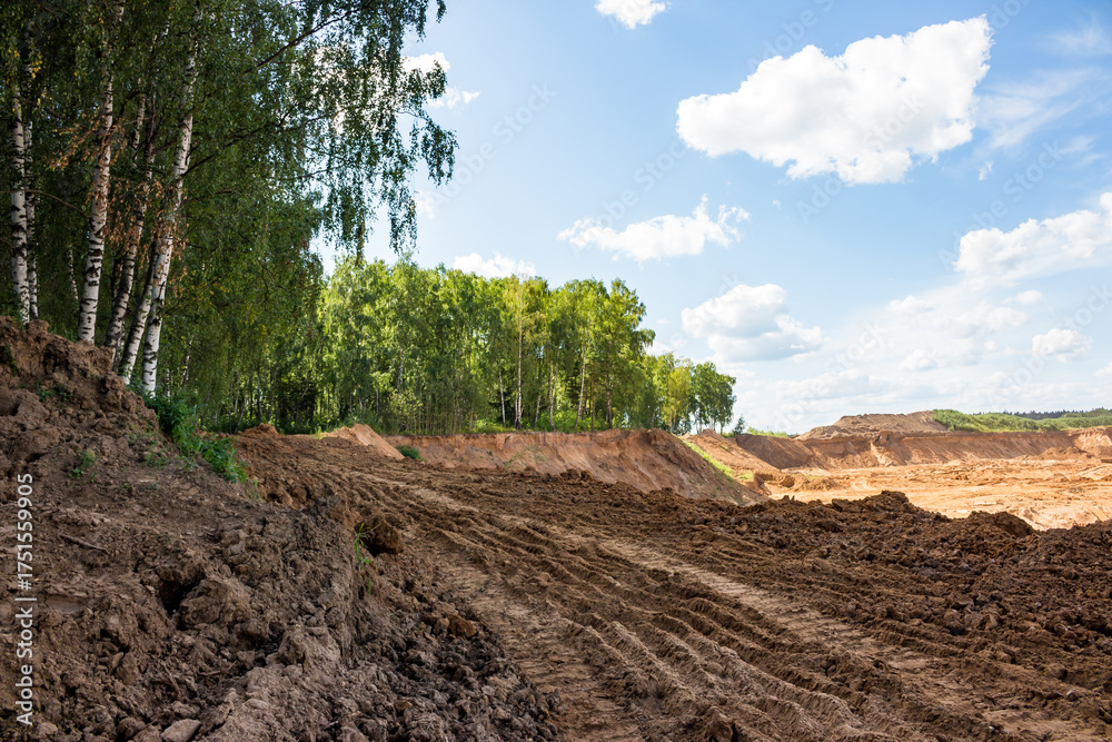 Fototapeta premium Rough brown soil with deep tire tracks dominates the foreground, hinting at ongoing excavation near a vibrant green forest and distant sand quarry under a clear blue sky
