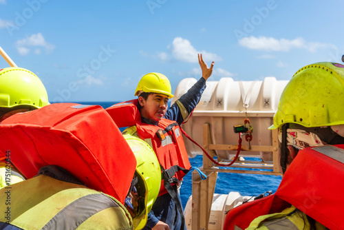 Deck officer wearing a yellow hard hat and orange life jacket demonstrates life raft deployment on a merchant ship. 