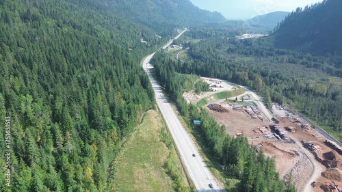 Cinematic drone footage captures traffic moving along a highway in a remote wilderness. The scene shows infrastructure development and a large land clearing project.