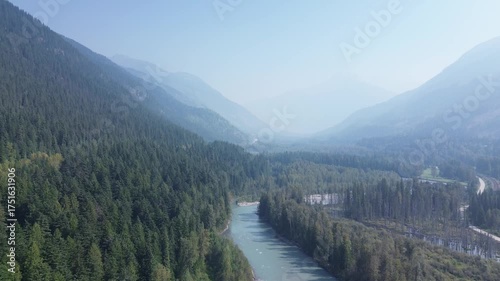 Cinematic drone footage captures the majestic scale of a remote mountain pass. A clear river snakes through the dense forest, leading the eye into the misty, layered peaks beyond.