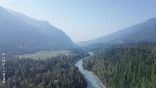 Cinematic drone footage captures the majestic scale of a remote mountain pass. A clear river snakes through the dense forest, leading the eye into the misty, layered peaks beyond.