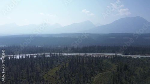 Cinematic drone footage reveals a breathtaking, layered landscape. A wide river flows through an endless forest, with distant, misty mountain peaks creating an epic backdrop.