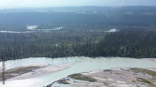 Cinematic drone footage captures a vast wilderness under a hazy sky. A milky, glacial river flows through the forest, while distant smoke indicates active wildfires.