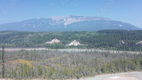Cinematic drone footage over a rugged landscape. A glacial river flows through a forest containing industrial piles, highlighting the impact of human activity on the wilderness.