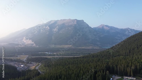 Cinematic drone flight over a vast pine forest, revealing a large mining or factory operation. The scene contrasts heavy industry with the pristine wilderness of the surrounding mountains.