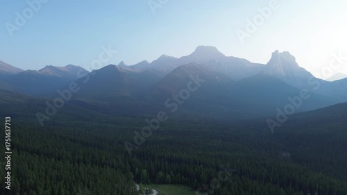 Cinematic drone footage flies over a dark, moody forest towards a breathtaking mountain vista at sunrise. The sun's rays create a mystical haze over the rugged, silhouetted peaks.