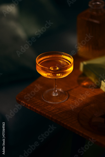 Close-up of an elegant cocktail glass filled with an amber drink, bathed in warm evening light on a wooden table. A cozy and sophisticated still life scene.