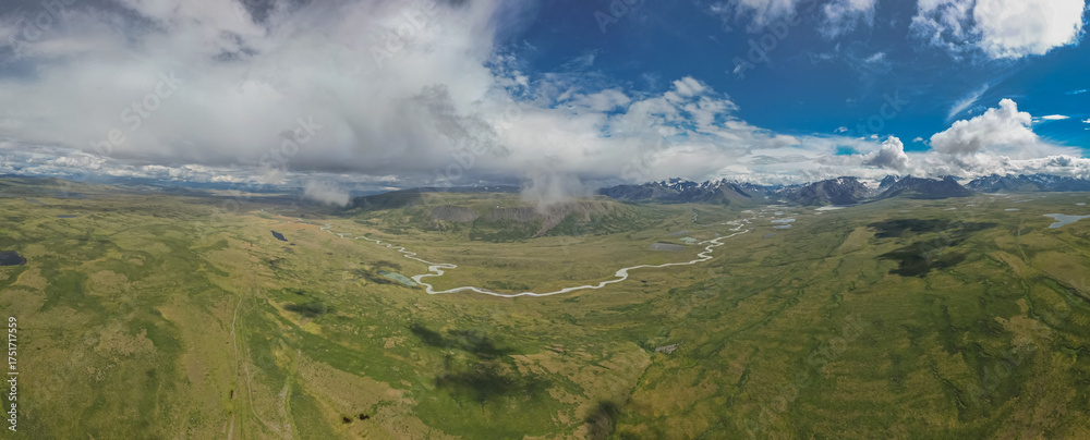 Fototapeta premium A panoramic photo taken from a drone shows snow-covered mountains in the background, green fields below, and a white river flowing through the meadows and fields.