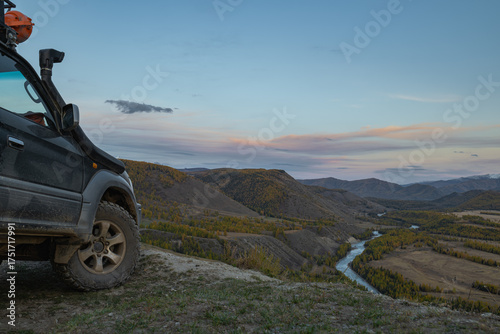An off-road vehicle with mud tires is parked high on a mountain, showcasing an off-road tire. An off-road vehicle is parked on a cliff, overlooking a forest and a river in the distance.